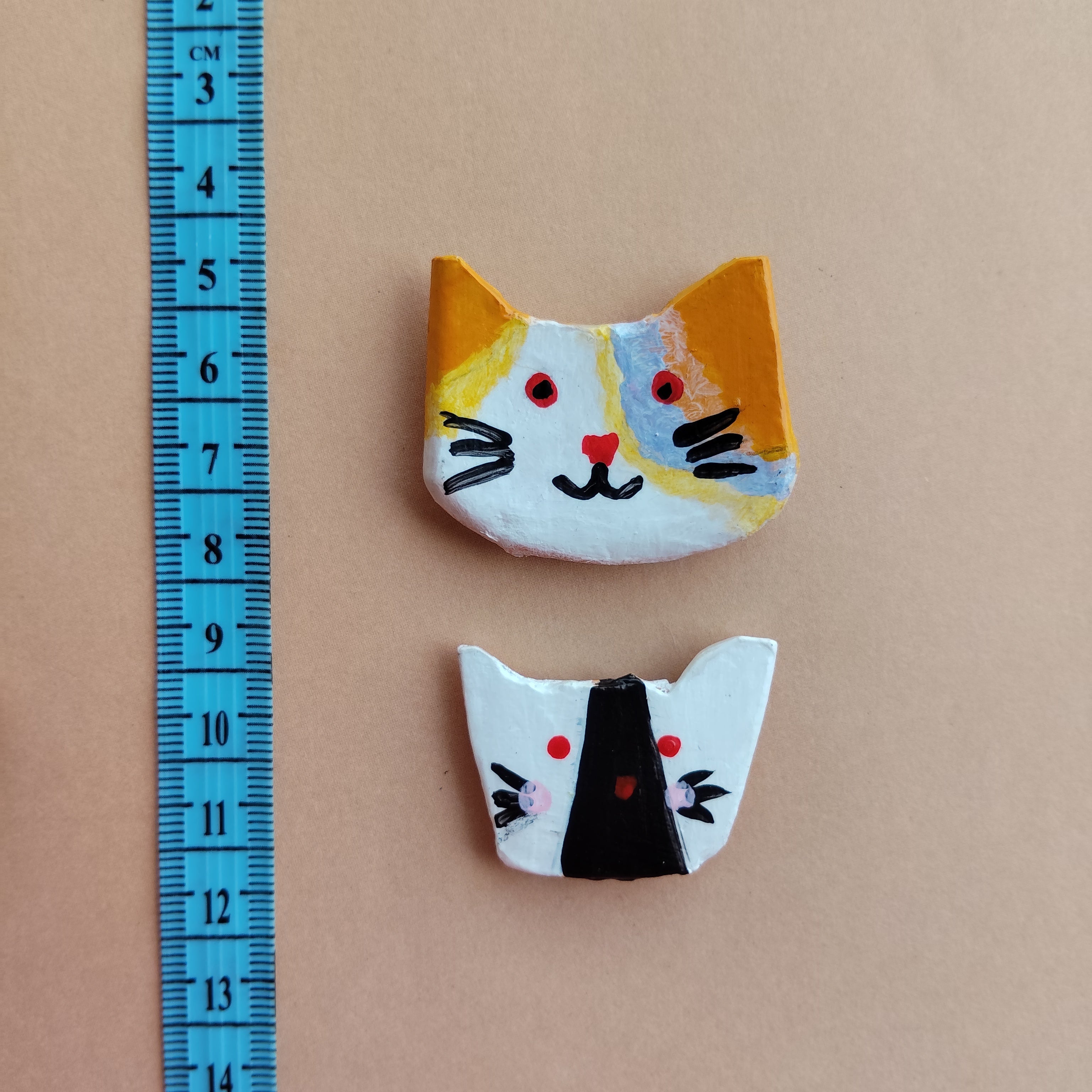Two handmade cat face with artistic designs on a beige background, accompanied by a blue ruler for scale. - Sustainable Home Decor & Upcycled Glass