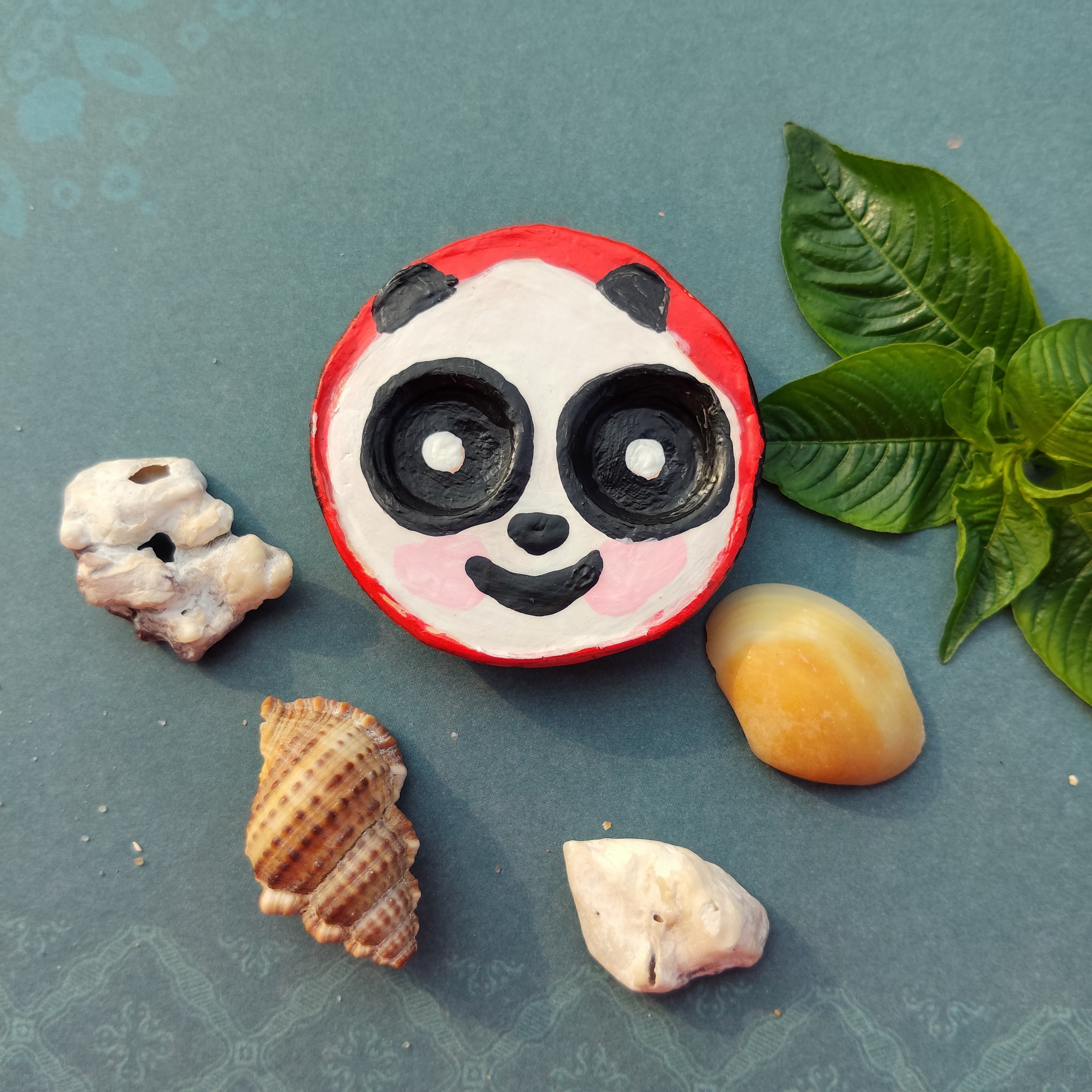 Decorative stone with round panda design surrounded by shells and a leaf on a textured surface. - Sustainable Home Decor & Upcycled Glass