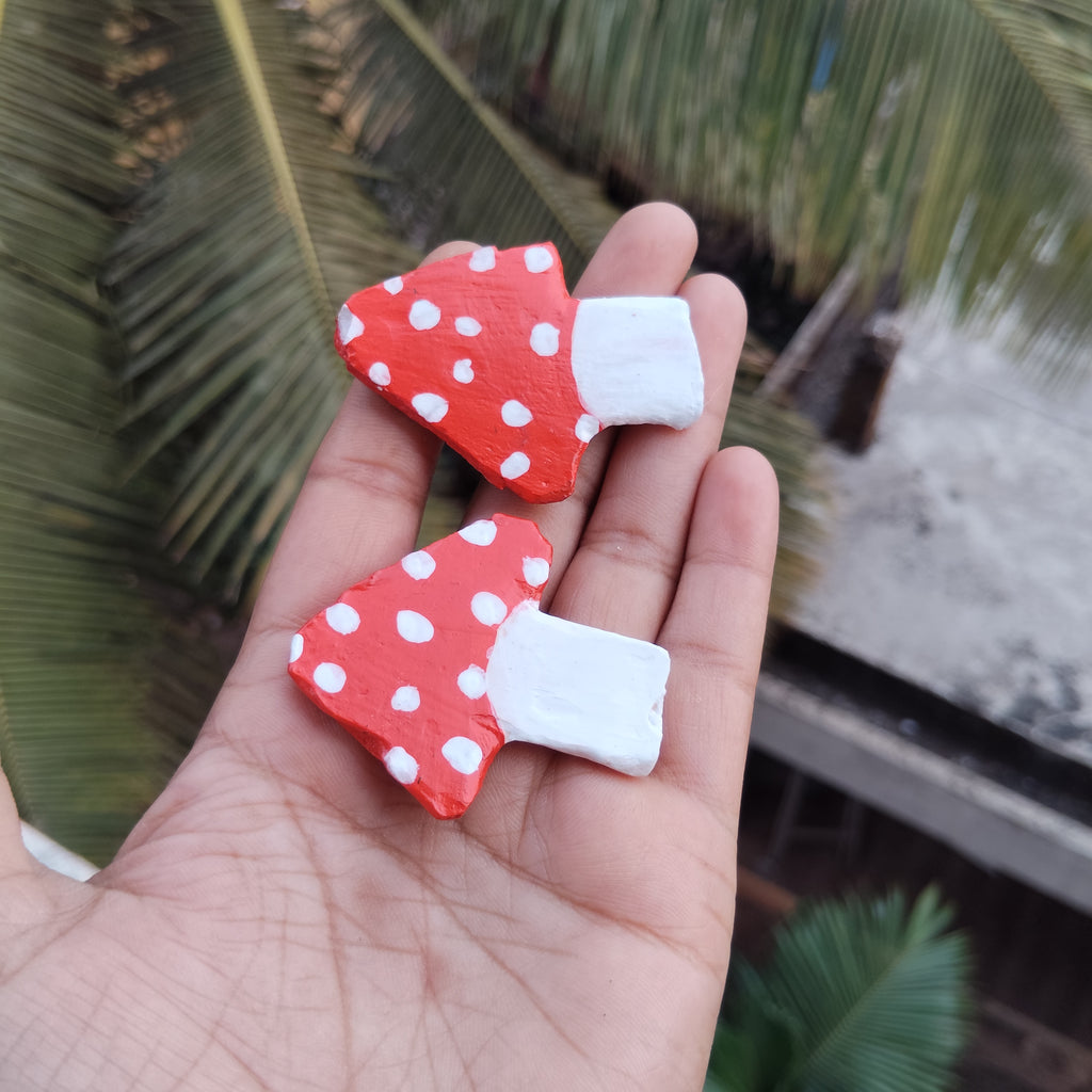 Two red polka dot mushroom-shaped magnets held in a hand with a blurred green leafy background. - Sustainable Home Decor