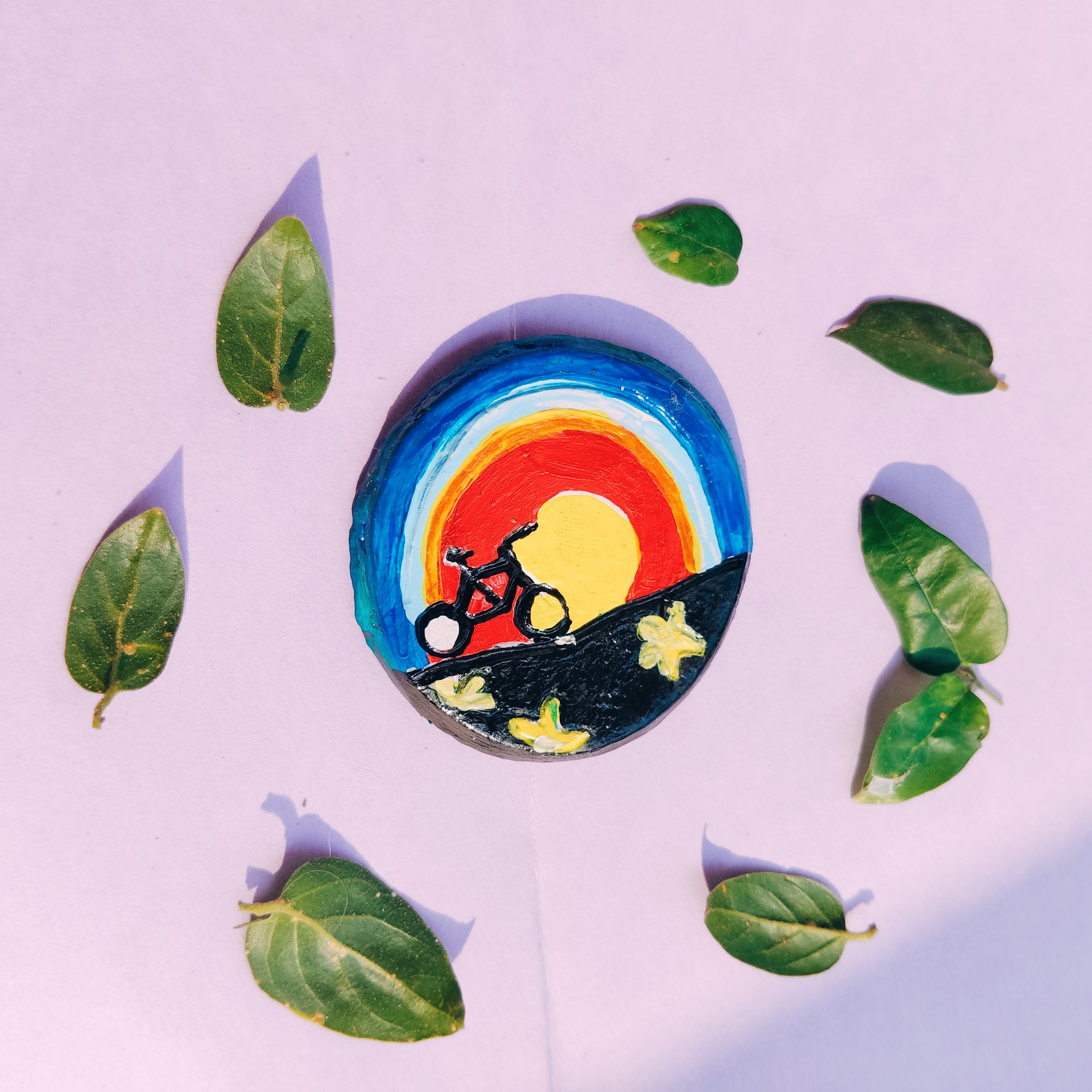 Colorful circular design  hand-painted on a clay surface with a bicycle silhouette, surrounded by green leaves on a light purple background. Its a fridge magnet.
