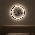 Round 3d textured artistic wall light fixture resembling a moon on a hanged on a dark wall above a bed.