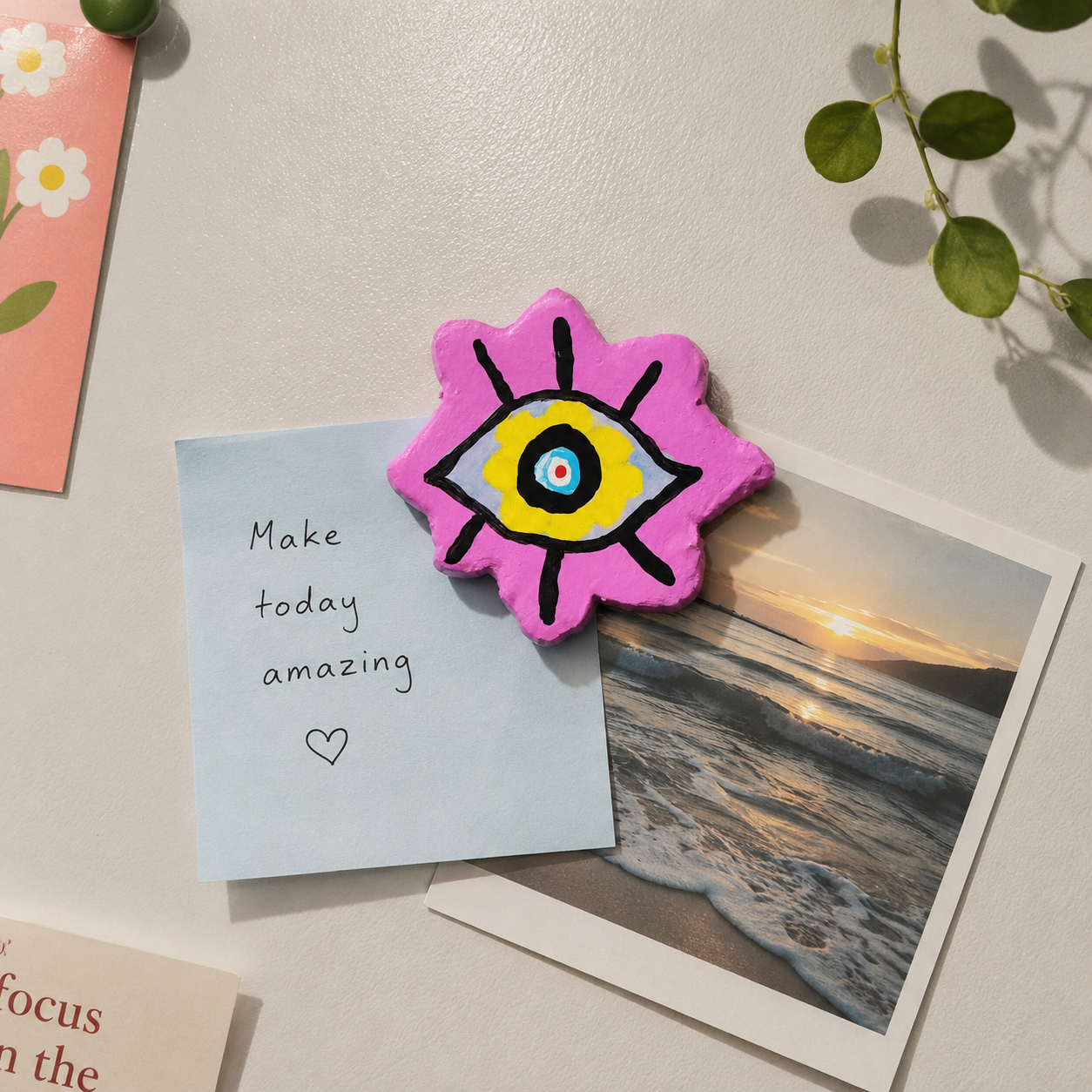 Pink flower-shaped magnet with a black eye design on a surface with cards and a photo of a sunset. - Sustainable Home Decor & Upcycled Glass