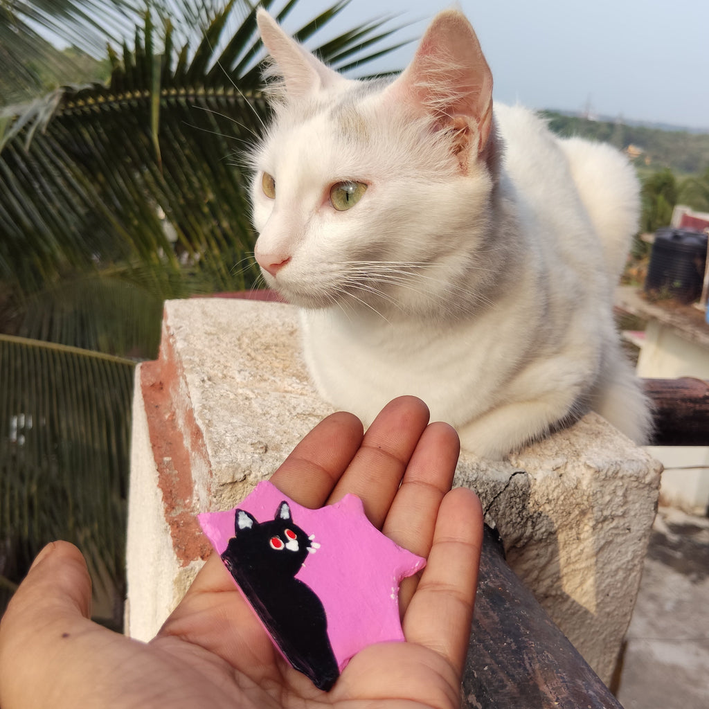 White cat sitting on a ledge with a hand holding a pink cute magnet with a black cat design. - Sustainable Home Decor