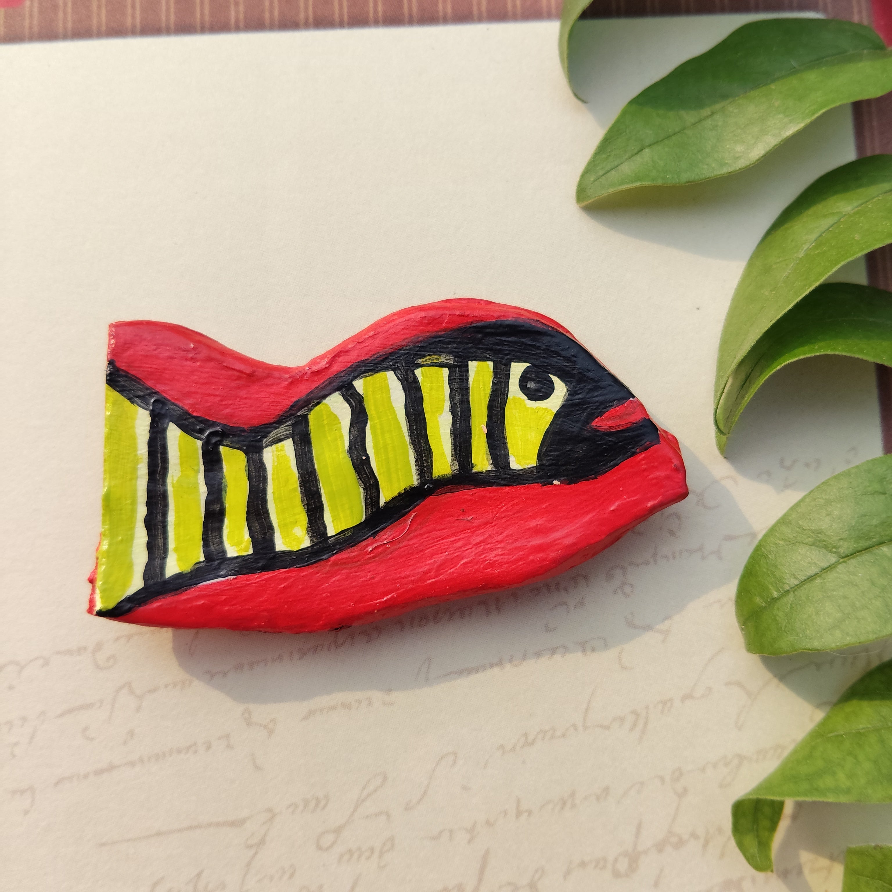 Decorative snake-shaped hand-crafted magnet with red, yellow, and black stripes on a textured surface with green leaves. - Sustainable Home Decor & Upcycled Glass