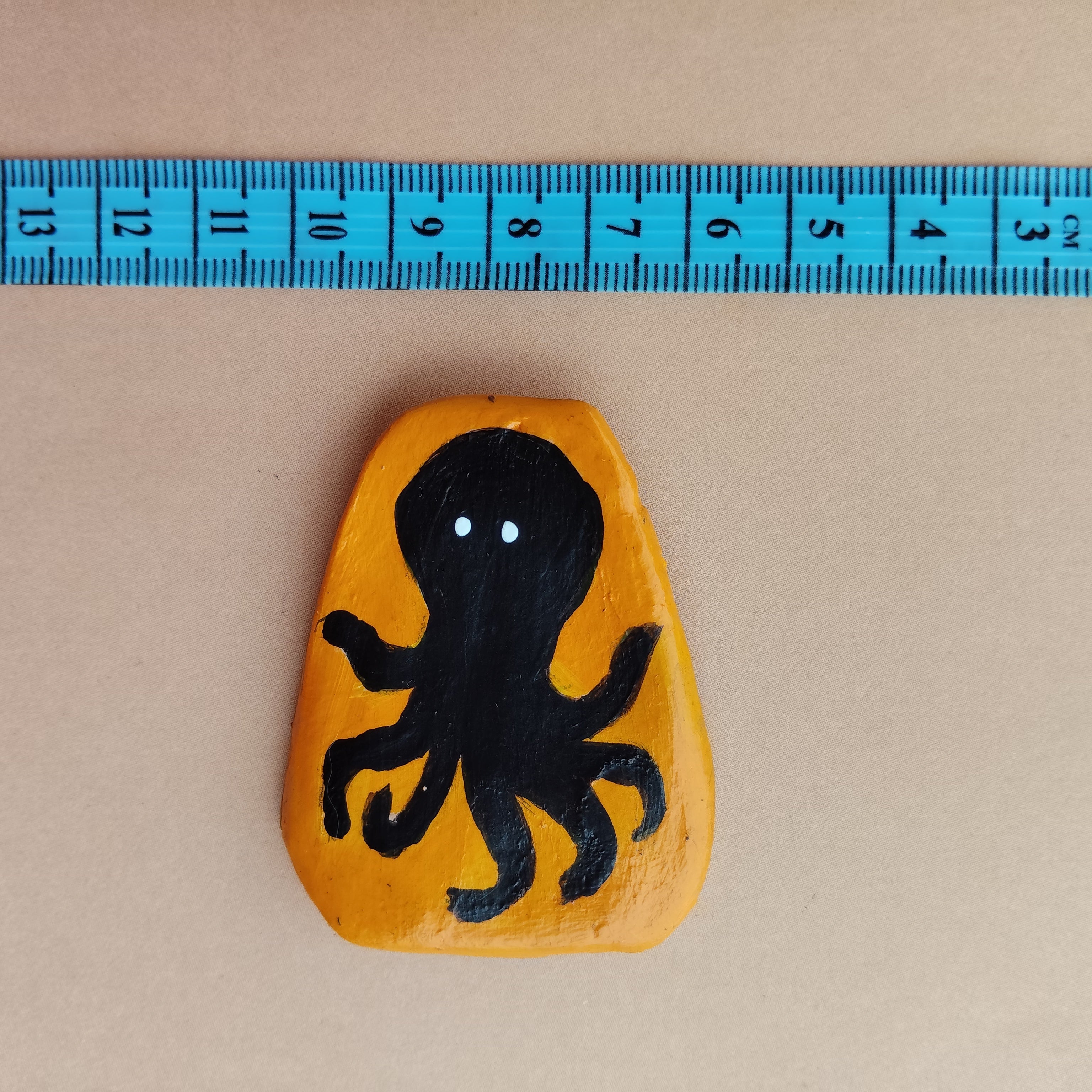Handcrafted magnet with a black octopus design on an mustard color background, measured against a blue ruler. - Sustainable Home Decor & Upcycled Glass