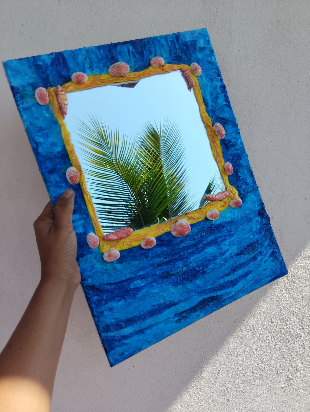 Hand holding a mirror with a colorful wavy textured frame in a blue background along with a functional mirror.