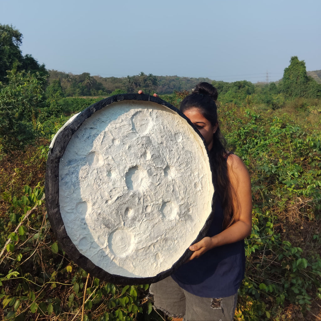 A girl holding a large circular hand-crafted 3d textured art resembling the moon in a natural setting