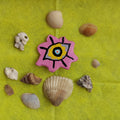 Pink handmade eye with a colorful floral design surrounded by seashells on a lemon yellow background.
