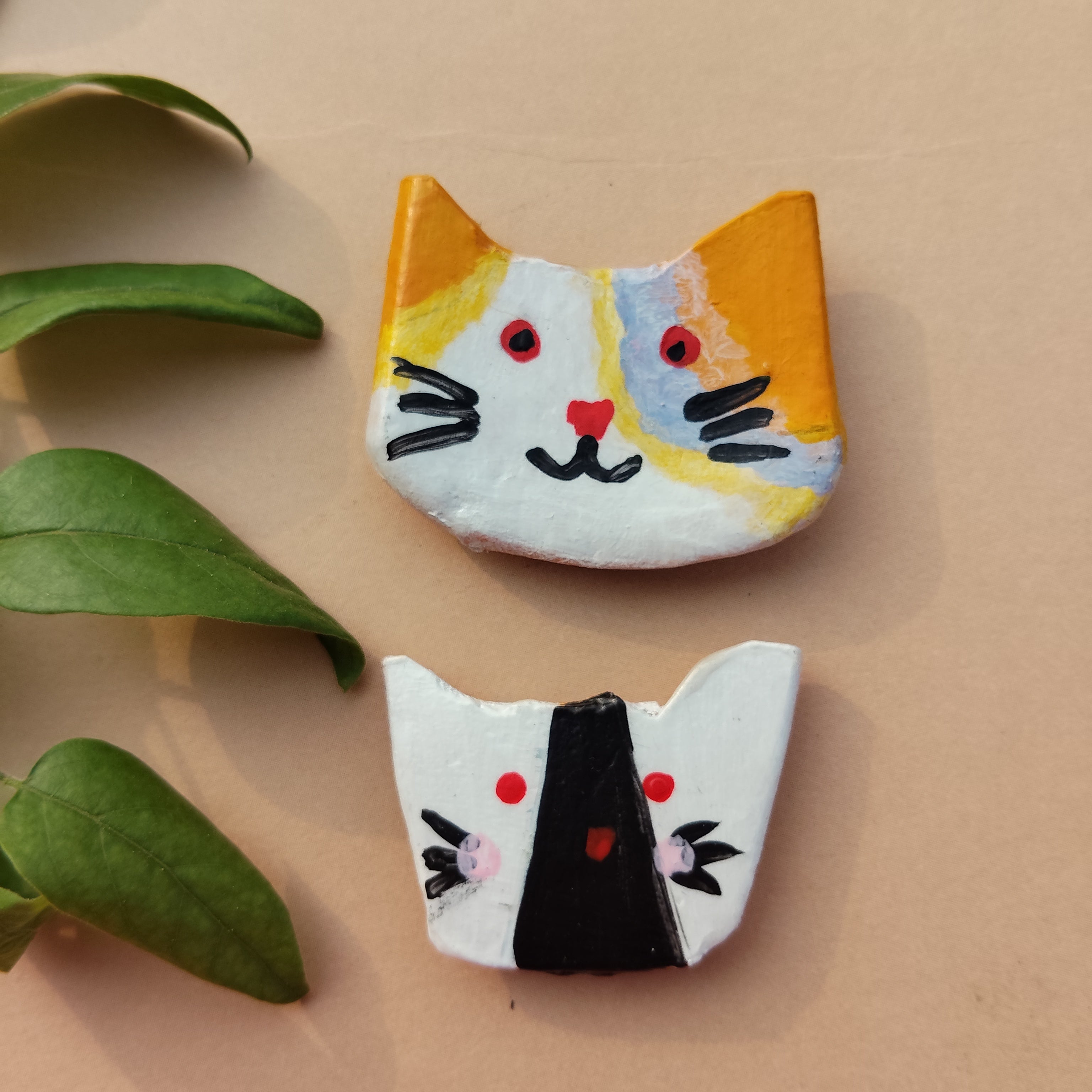 Handmade cat magnet pair  lying vertically on beige background with green leaves on sides. - Sustainable Home Decor & Upcycled Glass
