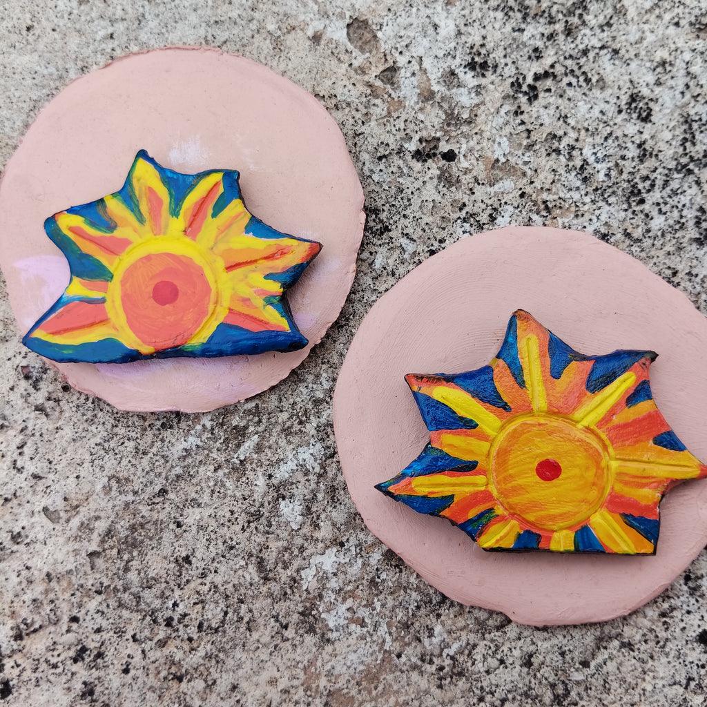Two colorful sun-shaped hand-painted magnets on pink bases against a textured surface - Sustainable Home Decor