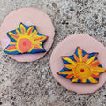 Two colorful sun-shaped hand-painted magnets on pink bases against a textured surface