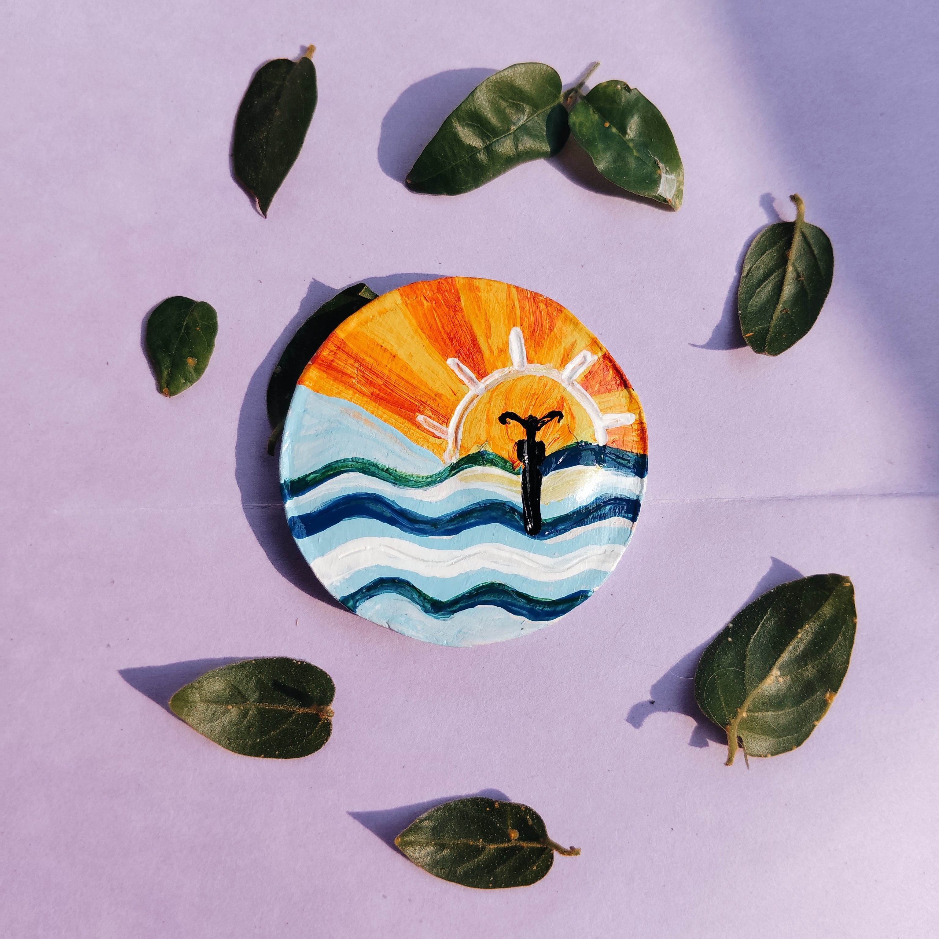 Circular artwork with a sun and wave design surrounded by live leaves on a purple background. It hand-crafted miniature magnet which is eco-friendly and pocket-friendly.